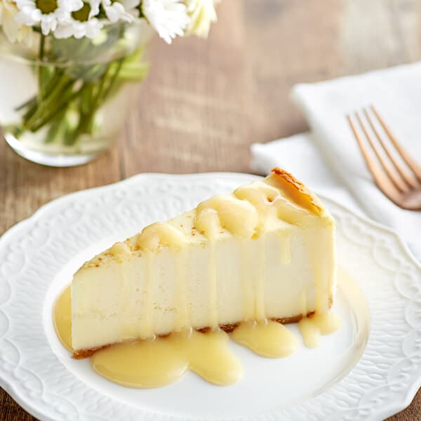A slice of cheesecake drizzled with white chocolate sauce on a white plate.